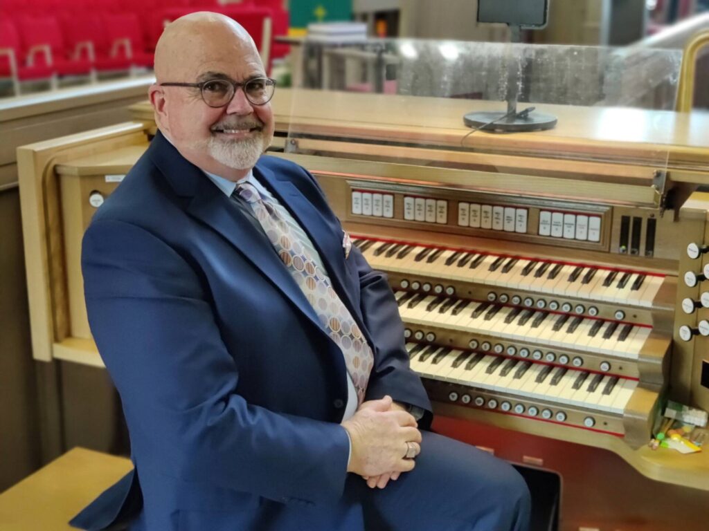 Man in suit seated beside a church organ console