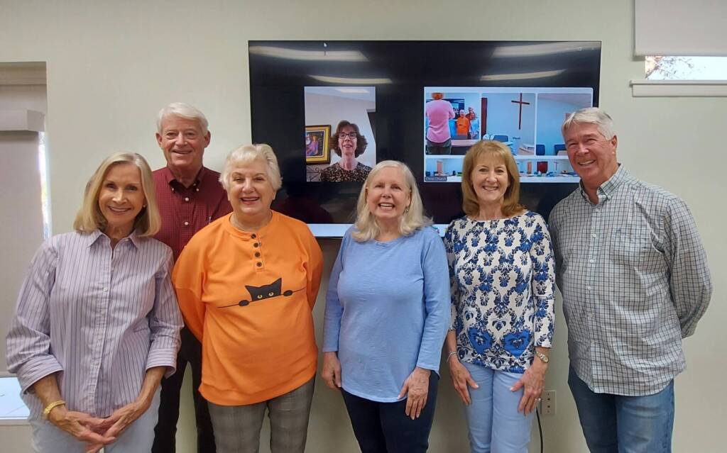 Six smiling adults pose beside church video call screen.