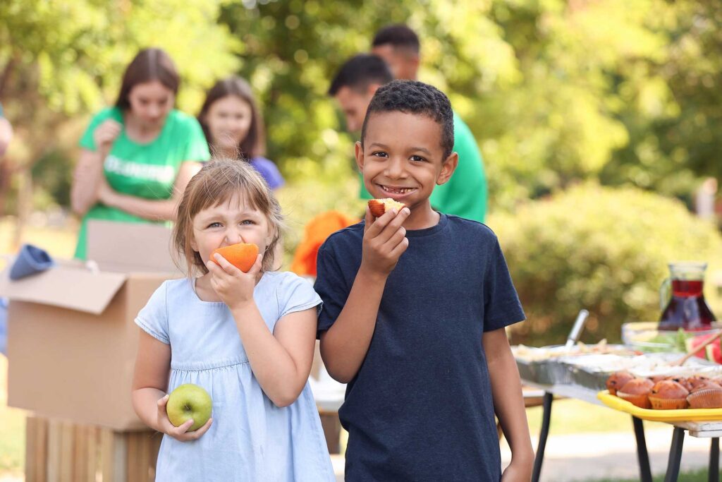 Two children eat fruit at an outdoor community event.